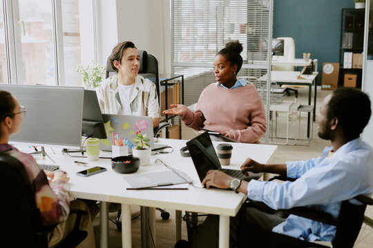 Biracial colleagues discussing next goals while sitting at white table in modern IT company office equipped with electronic devices