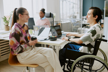 Young Caucasian female colleague holding cup of coffee and chatting lovely with her Caucasian male colleague with disability