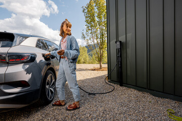 Young woman plugs a charger into electric vehicle standing with smart phone near her house outdoors. Concept of modern technologies, EV cars and sustainable lifestyle