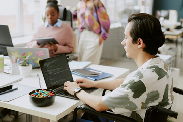 Fototapeta premium High angle shot of young IT specialist with disability writing code while his female coworkers working in blurred background