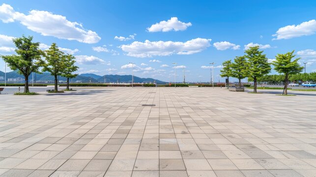 A wide, empty city square with a paved surface and trees lining the edges. The sky is bright blue with fluffy white clouds