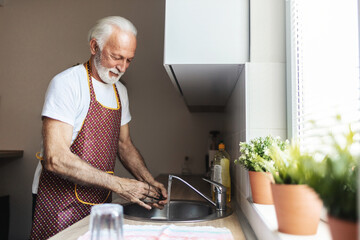 Senior white man standing next to a kitchen sink and rinsing a glass with running water inside a kitchen.