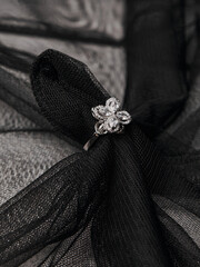 A silver ring with a floral pattern and precious stones rests on a black textured fabric, creating an elegant contrast.