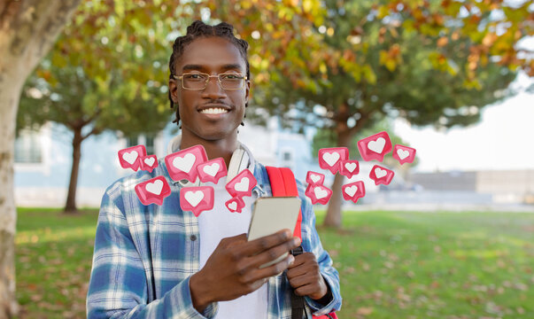 A young black man wearing a plaid shirt and glasses stands in a park with a backpack, smiling as he checks phone. Digital hearts and notifications float around him, suggesting social media activity.