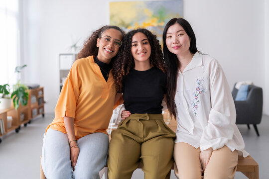 Three diverse women smile together in a bright modern office The women are dressed in colorful outfits and are standing close to each other showcasing their camaraderie and friendship