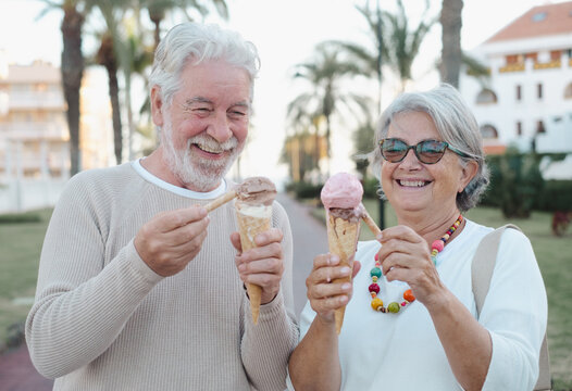 Smiling retired couple having fun eating ice cream cone in the park. Joyful elderly lifestyle concept. Two white-haired seniors enjoying a sweet fresh dessert