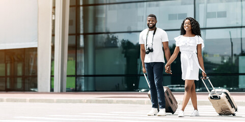 Fototapeta premium A smiling black couple walks together outside of an airport building, pulling their luggage behind them. They are both looking forward and appear to be enjoying their time together.