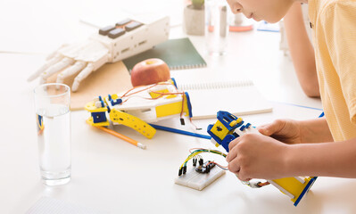 A young student sits at a desk, focusing intently on building a robotic hand. The hand is a bright yellow and blue, and the student is holding it with both hands, studying the wires and circuitry