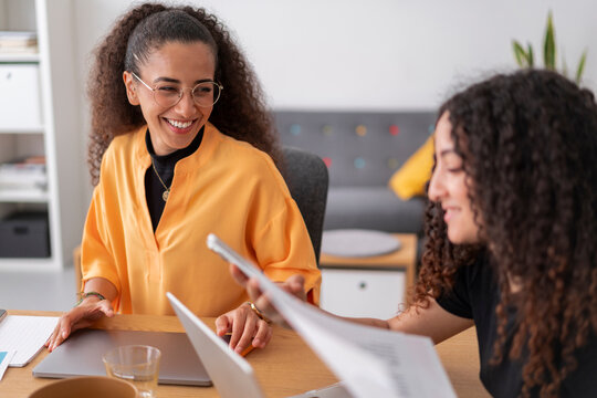 Two women are collaborating in a bright and modern office. One is holding a document, and the other is smiling and looking at her. They are sitting at a wooden desk with laptops and office supplies.