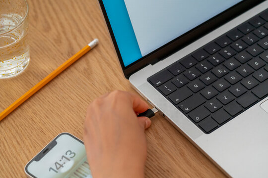 A close-up of a hand plugging a USB cable into a laptop, alongside a smartphone and a glass of water. The image highlights the integration of technology in everyday work tasks.