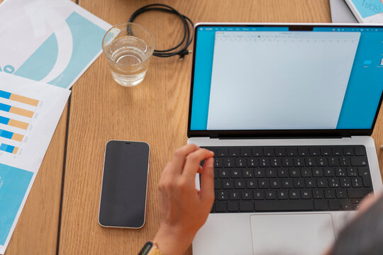 A young professional typing on a laptop while analyzing charts and graphs during a productive teamwork session in a modern office setting, surrounded by documents and notes