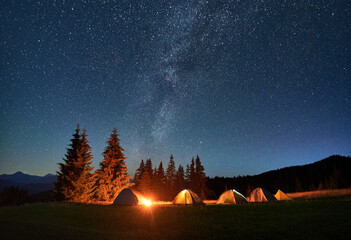 Night camping in mountains under starry sky. Tourist tents in campsite near burning campfire under...