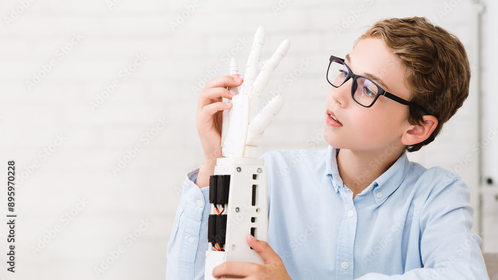 Poster A young boy wearing glasses examines a robotic hand in a classroom setting. He appears to be intrigued and fascinated by the technology, demonstrating the growing interest in robotics