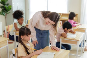 A teacher assists a young student in a classroom setting, while other students work at their desks.