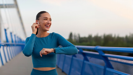 Beautiful female jogger listening to music in earbuds and wearing smartwatch while running on...