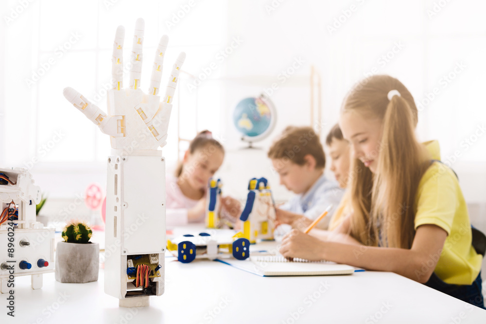 Aufkleber A robotic hand model stands in the foreground of the image, in focus, while a group of children work on a project in the background. The classroom is bright and filled with natural light.