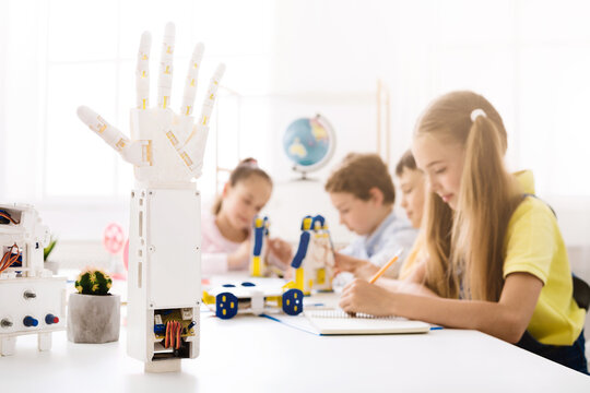 A robotic hand model stands in the foreground of the image, in focus, while a group of children work on a project in the background. The classroom is bright and filled with natural light. - Powered by Adobe