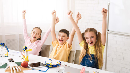 Three children, two girls and one boy, sit at a table with their arms raised in victory. They have completed a successful STEM project and are beaming with pride.
