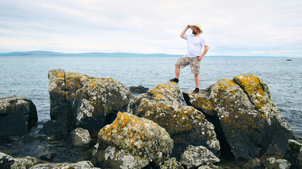 Handsome bearded man with straw hat standing on the rocks by the ocean at wild Atlantic way, adventure and lifestyle concept, summer, vacation, nature background