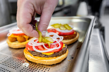 Hand placing a pickle on a burger with onions, tomatoes, and lettuce