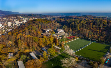 Aerial view of autumn forest, soccer field, and school surrounded by mountains and hills, Biel, Bern, Switzerland.