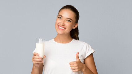 Dairy Products Concept. Woman Holding Glass Of Milk Gesturing Thumbs Up Approving Taste Standing In Studio Over Gray Background. Panorama