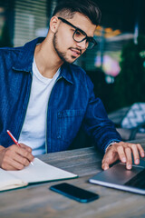 Young caucasian male student in spectacles learning online course via laptop computer  and making...