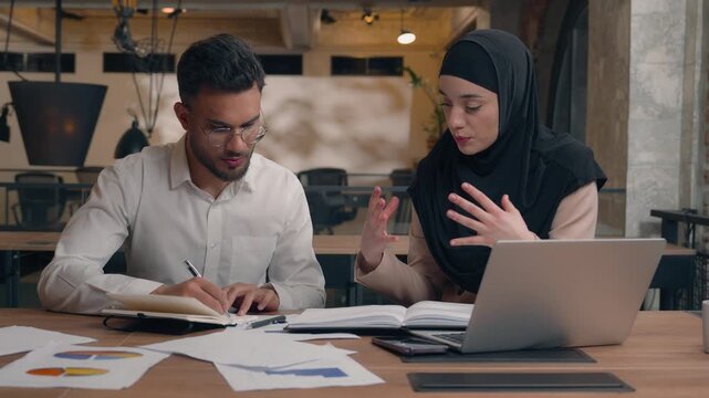 Two business colleagues muslim islamic businesswoman in hijab Arabian businessman working with laptop together in office woman coach talk consult explain teach advice trainee man writing in notebook