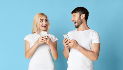 A young woman and man are standing in front of a blue background, both holding smartphones. The woman is looking at the man's phone with a surprised expression