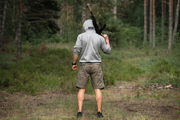 A man in shorts and a hooded sweatshirt in the forest with an assault rifle in the forest, viewed...