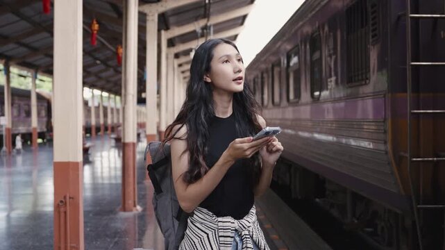 Young woman with a backpack uses her smartphone for navigation at a train station, looking around for directions and appearing confused