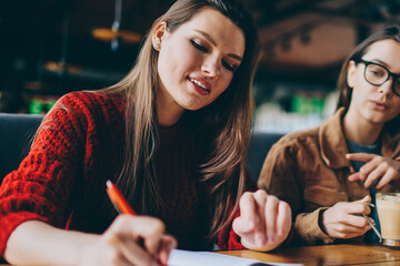 Positive carefree hipster girl writing essay with excited idea in textbook sitting near female friend with caffeine beverage, two happy successful women studying together at university cafeteria