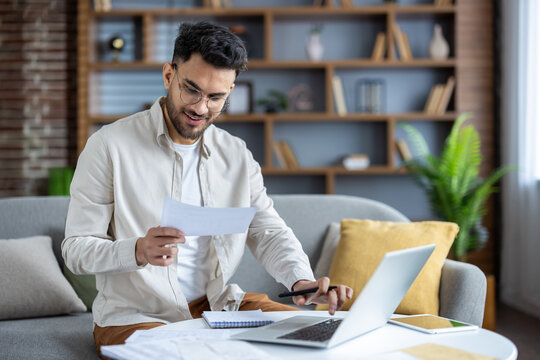 Young man working from home with laptop, reviewing documents and smiling - Powered by Adobe