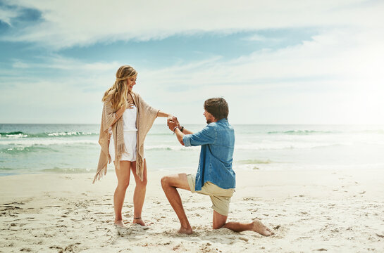 Marriage, proposal and couple with love at beach for engagement, happiness and surprise on vacation. Blue sky, waves and man with question on knee for romantic gesture, support and holiday in Venice