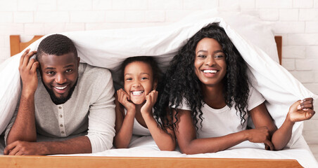 Cheerful black mother, father and daughter hiding under blanket, playing on bed at home, copy space