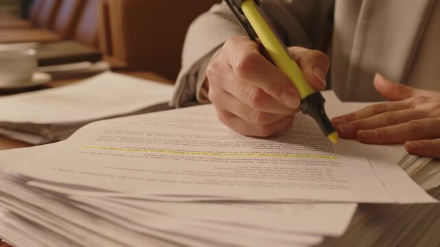 Close-up view of unrecognisable business person highlighting lines in document on top of pile of papers using yellow textmarker