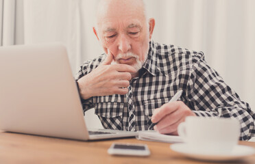 Mature man using laptop and writing in notepad at home desk. Education, recipe concept