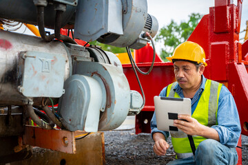 A man in a yellow safety vest is looking at a tablet while standing next to a machine. He is wearing a hard hat and he is focused on his work