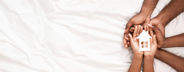 Adoption and family housing concept. Cutout paper house in hands of african american family over white bed sheet background. Closeup, panorama