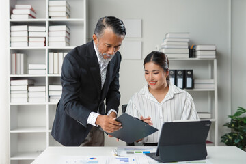 Experienced executive with a clipboard is discussing a business plan with a junior coworker using a tablet and paperwork