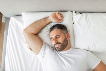 A man with a beard is sleeping soundly in a white bed. His arm is extended above his head, resting on a pillow. The man is wearing a white shirt and his eyes are closed.