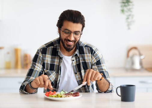 Positive arab guy hipster having healthy breakfast at home, eating fresh vegetables, sausages, drinking coffee, sitting at kitchen table, copy space. Handsome indian man starting day with nice meal