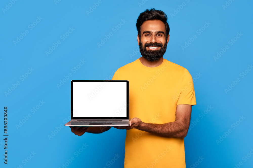 Poster a bearded indian man in a yellow t-shirt smiles while holding a laptop with a blank screen in front 