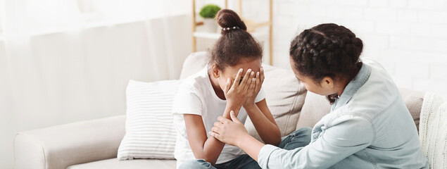 Teenaged african american girl comforting her crying little sister, sitting on sofa at home