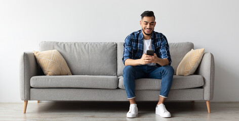 Cool Gadget And Application. Portrait of young smiling Arab man holding mobile phone, typing sms message, sitting on the couch in living room. Guy browsing internet, surfing web, using app, free space