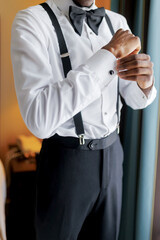 A dark-skinned man puts on a white shirt and buttons the cuff close-up. Stylish groom puts on a white shirt and suspenders while preparing for the wedding ceremony