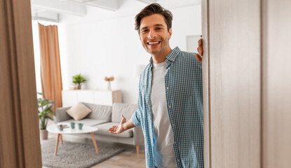 Portrait of cheerful man welcoming inviting visitor to enter his home, happy young guy standing in...