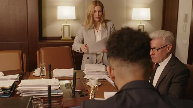 Female employee bringing stack of documents to conference room and putting it on wooden table, then giving several sheets to male boss who passing them to male colleague