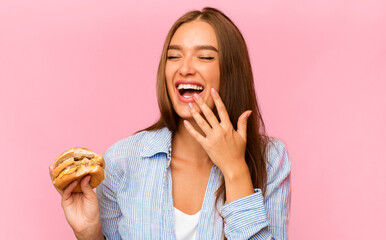 Junk Food. Young Woman Eating Burger And Laughing Standing Over Yellow Studio Background.