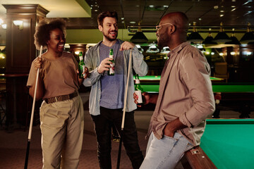 Happy young intercultural couple with beer and billiard cues standing in front of African American guy and discussing latest news